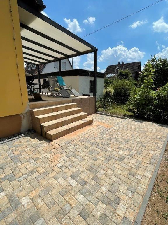 Überdachte Terrasse mit gelben Wänden, Treppe, zwei Liegestühlen; sonniger Tag, blauer Himmel.