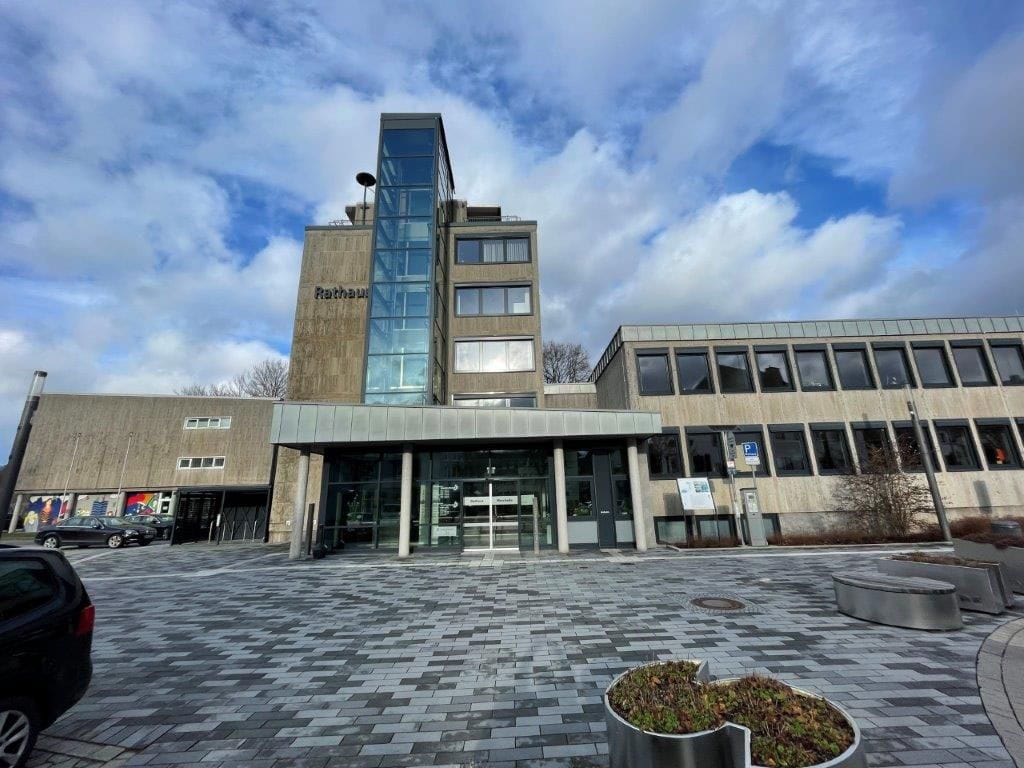 Ansicht eines modernen Rathauses mit Glasfassade, grauen Wänden und bewölktem Himmel im Hintergrund.